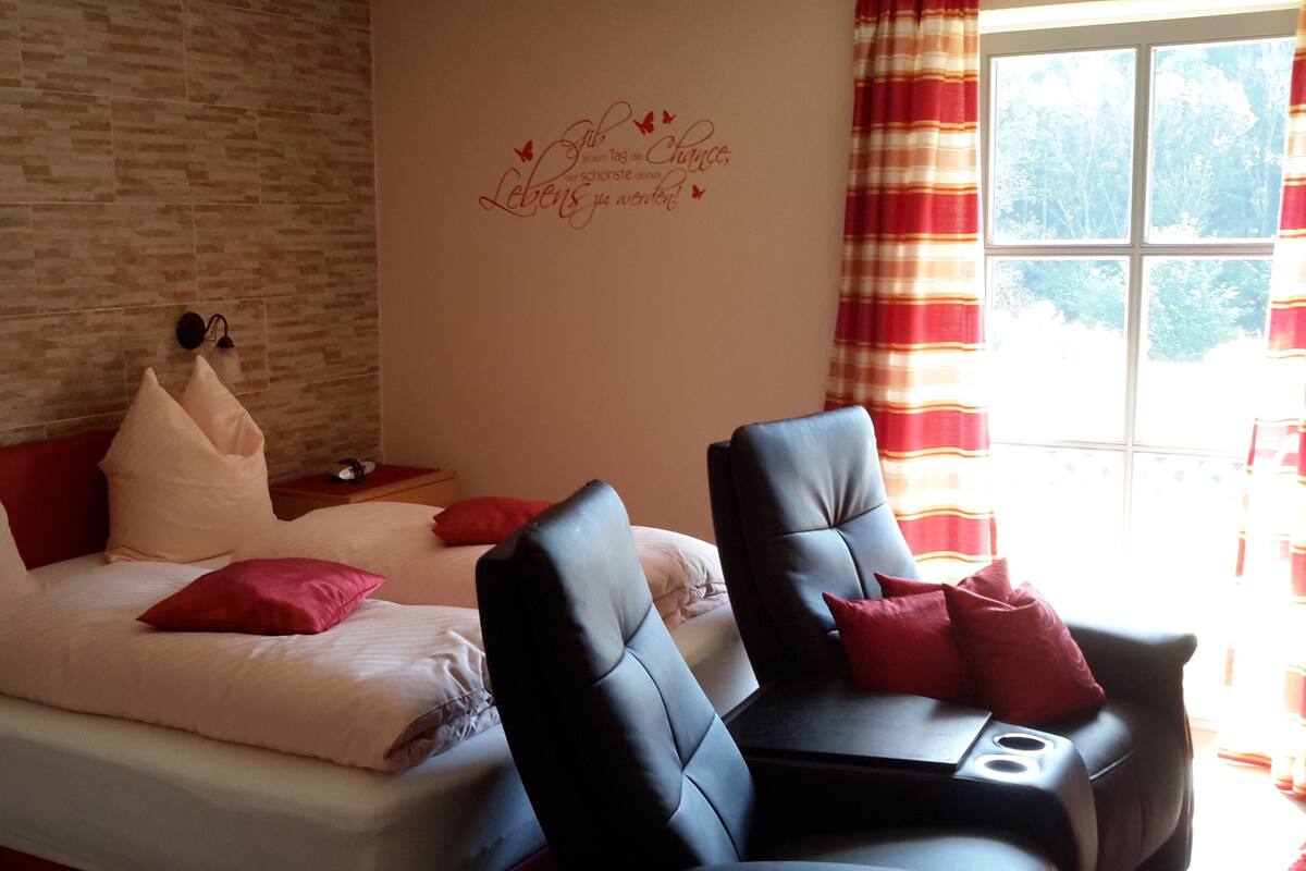 Une chambre à coucher avec un lit, deux chaises, une horloge murale et une fenêtre avec des rideaux.