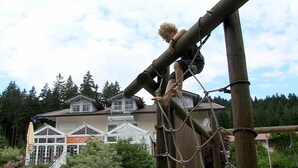 Une personne escalade une structure en bois à l’aide de cordes, avec des maisons et des arbres en arrière-plan.