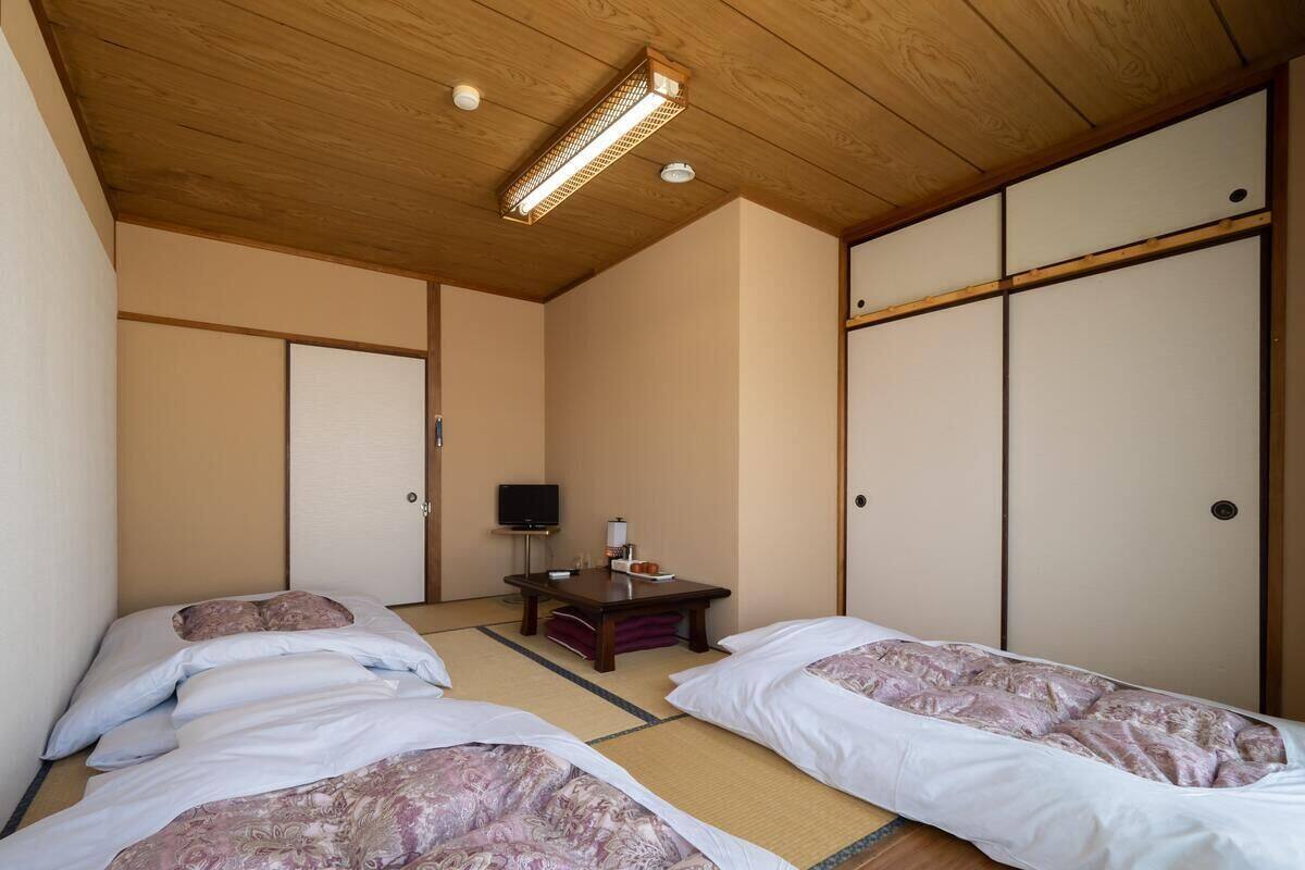 Een traditionele Japanse kamer met tatami vloeren, twee bedden met beddengoed met een patroon, een klein houten bureau met een televisie, en schuifdeuren.