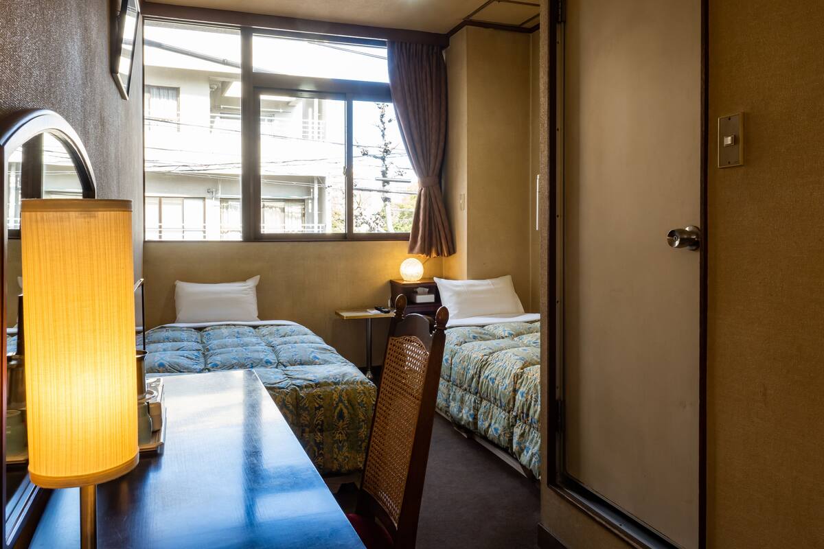 Een hotelkamer met twee bedden, een bureau, een lamp en een raam met gordijnen.