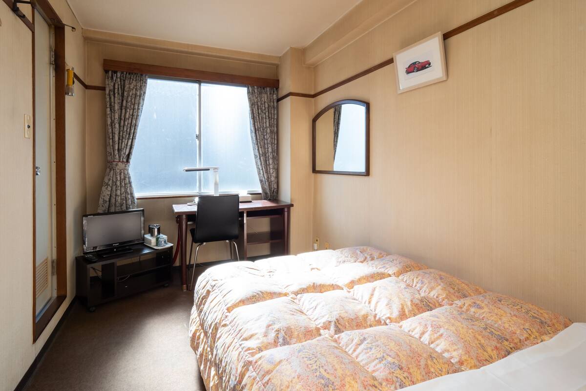 Een hotelkamer met een bed, een bureau met stoel, een computer en een raam met gordijnen.