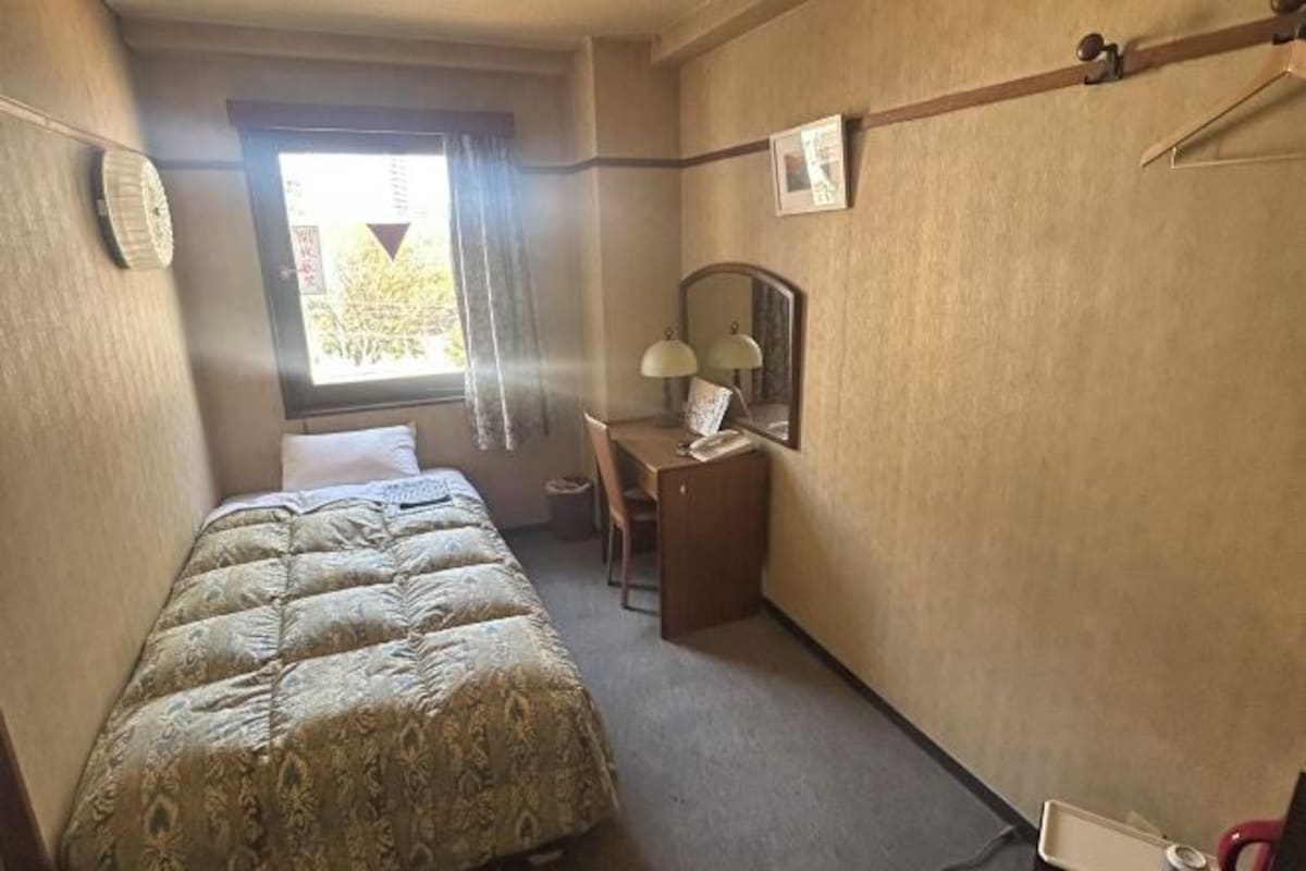 Une petite chambre d’hôtel avec un lit, un bureau, une chaise, un miroir et une fenêtre avec des rideaux.