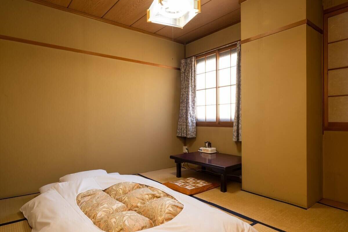 Een traditionele Japanse kamer met tatami vloerbedekking, een laag houten bed met witte beddengoed, een klein tafeltje met een theeservies en een raam met gordijn.