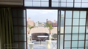 A hotel room with a bed, a small table with chairs, a view of the city through a sliding door, and a green curtain. - Business Hotel Nakayama (Nagoya)