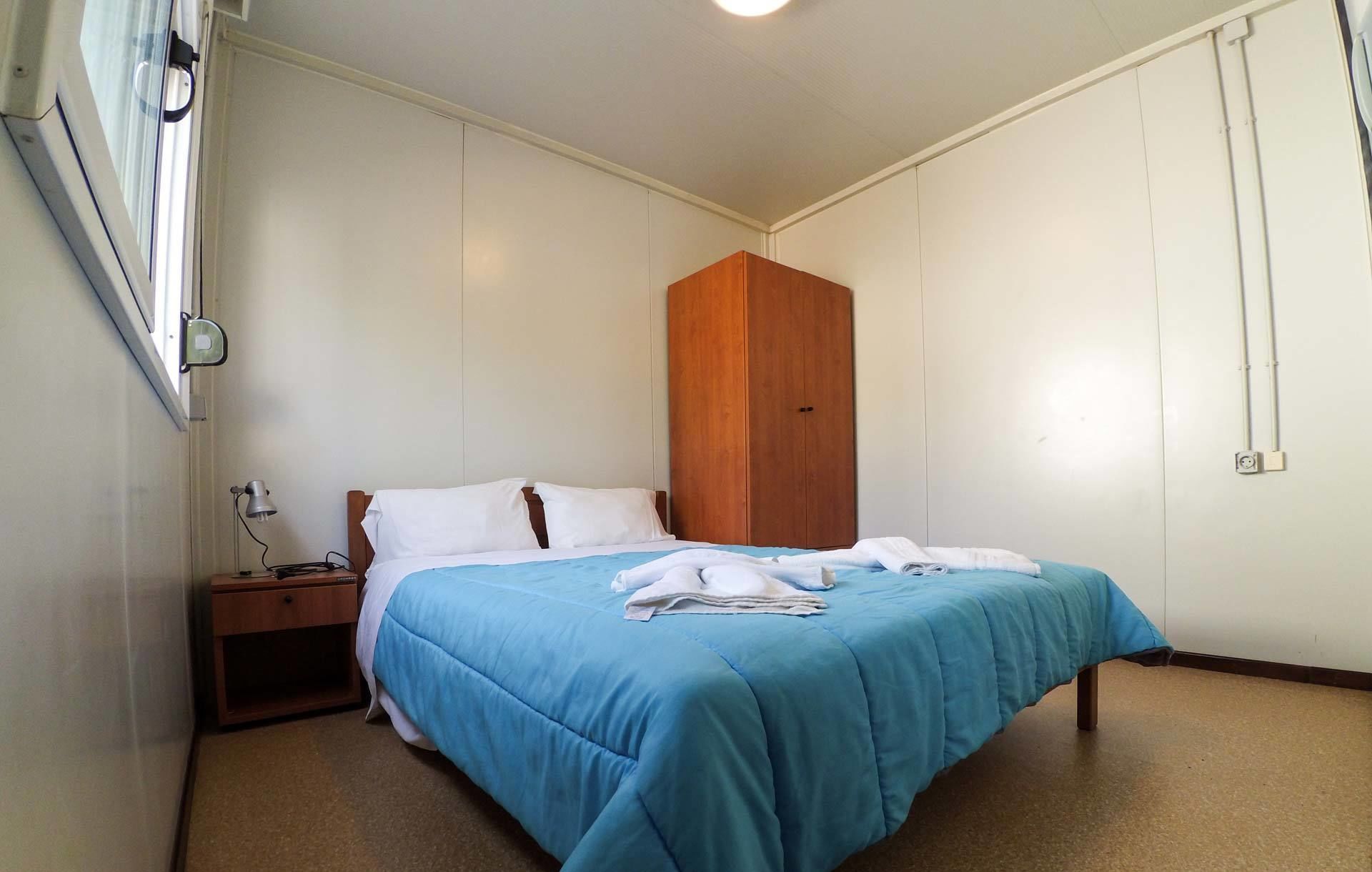 A bedroom with a bed, a wooden wardrobe, a nightstand, and a desk lamp.