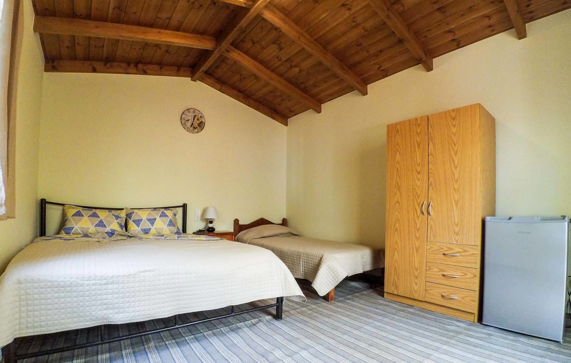 Un dormitorio con techo de madera, una cama con almohadas estampadas, una mesita de noche con lámpara, un armario y un refrigerador.