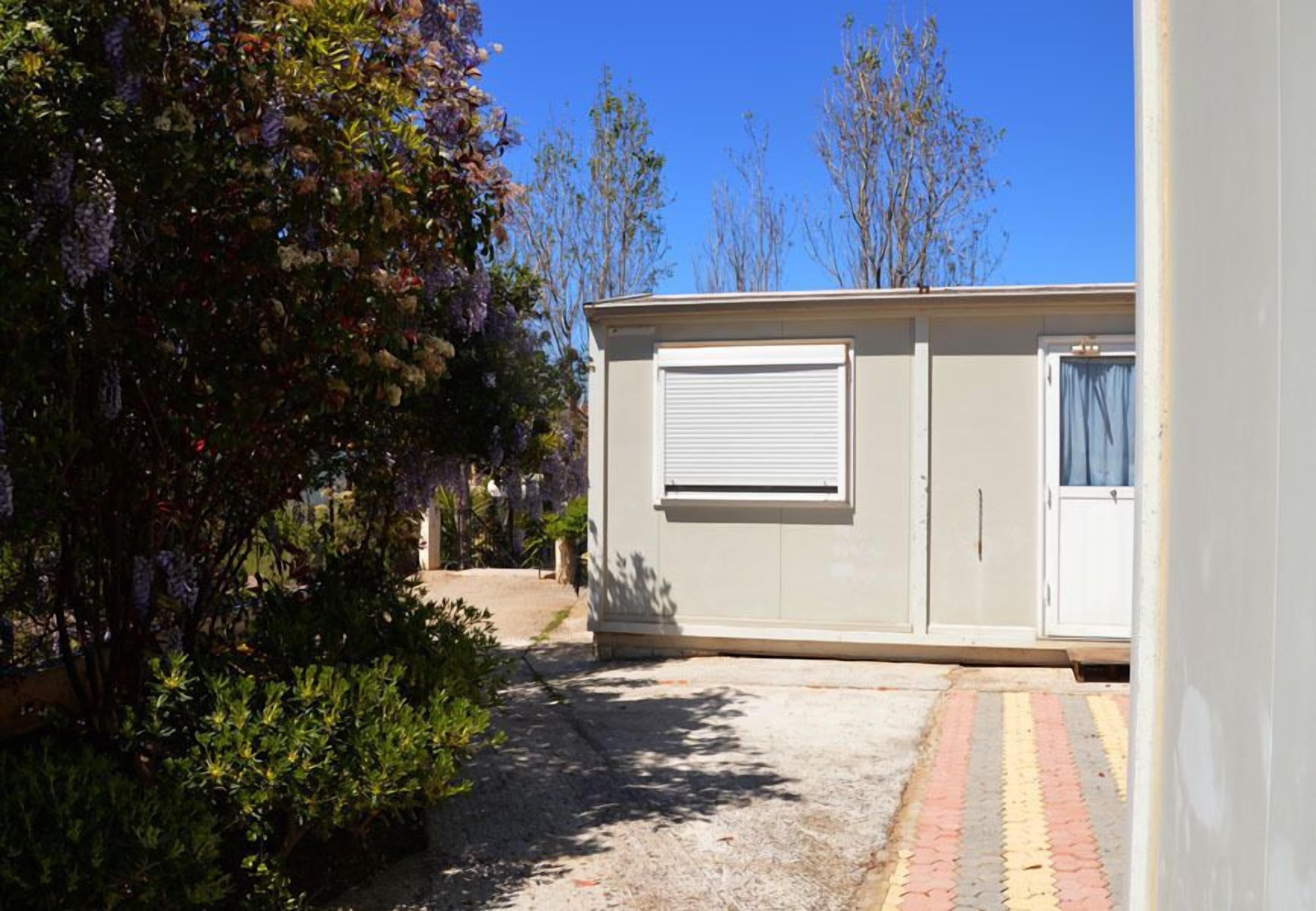 A small, single-story building with a closed shuttered window and a door, surrounded by dense foliage and a clear sky.