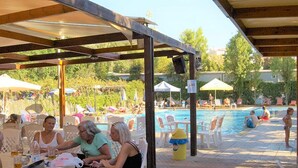 Un área de estar al aire libre con techo, mesas, sillas, piscina y sombrillas al fondo.