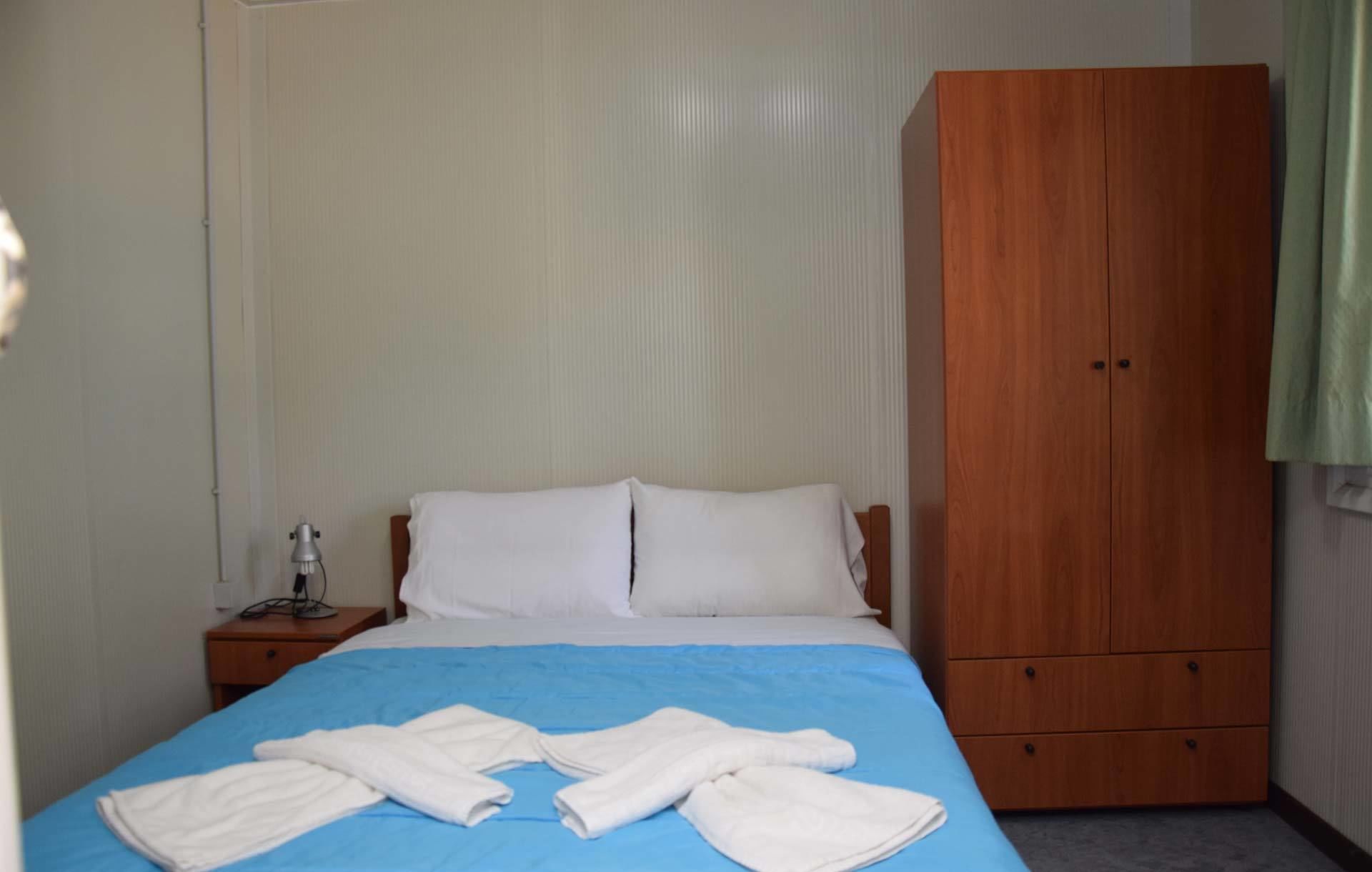 A hotel room with a bed, a wooden wardrobe, and a nightstand with a lamp.
