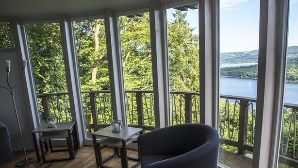 A room with large windows, a wooden deck, and a view of a lake and forest. - Holiday house "Waldhaus Anne" in Niedeggen - with a view of the Rursee (Nideggen)