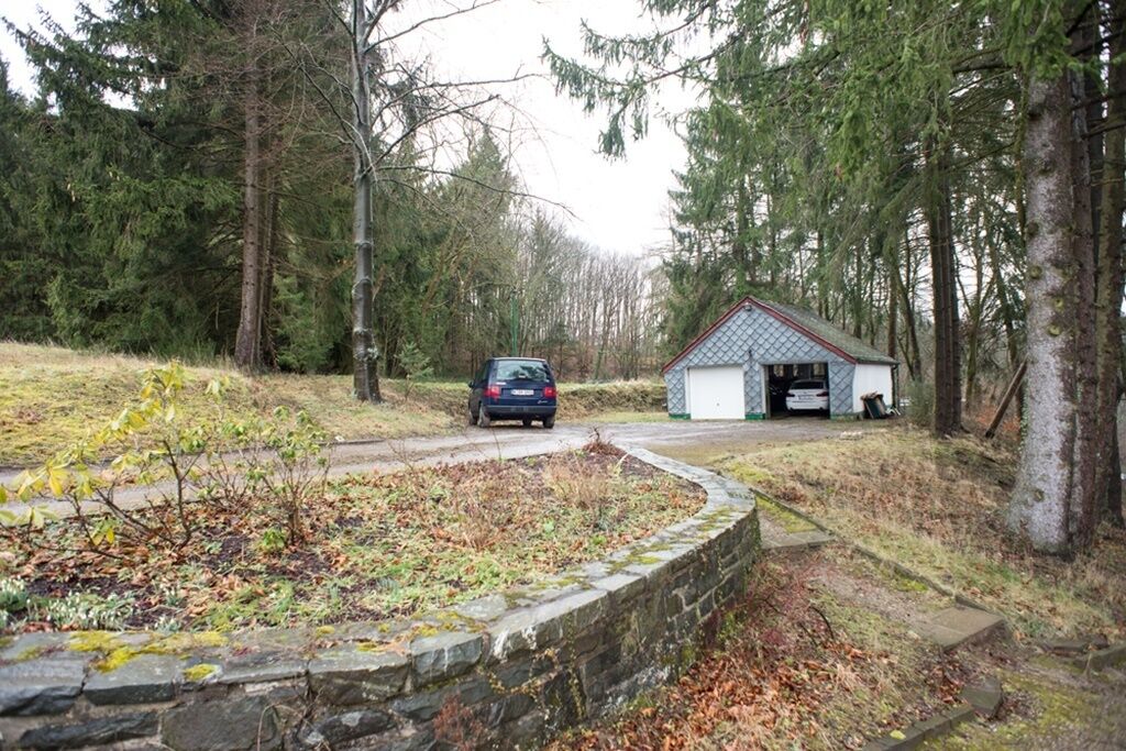 Un mur de pierre, une voiture et un bâtiment doté d’un toit en pente, le tout situé dans une zone boisée.