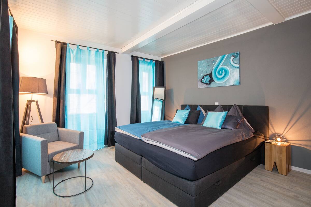 Quarto moderno com uma cama grande, uma cadeira, uma mesa de cabeceira e um quadro na parede.