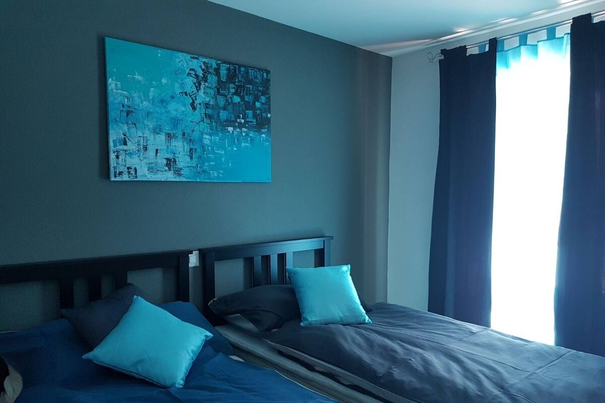 Quarto com cama, almofadas, cabeceira, um quadro na parede e janela com cortinas.
