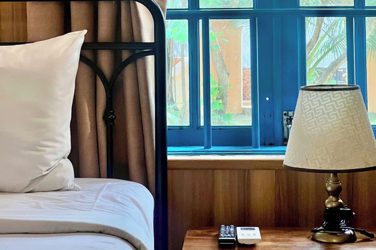 A room with a bed, a window with a view of greenery, a lamp, and a remote control on a wooden nightstand.