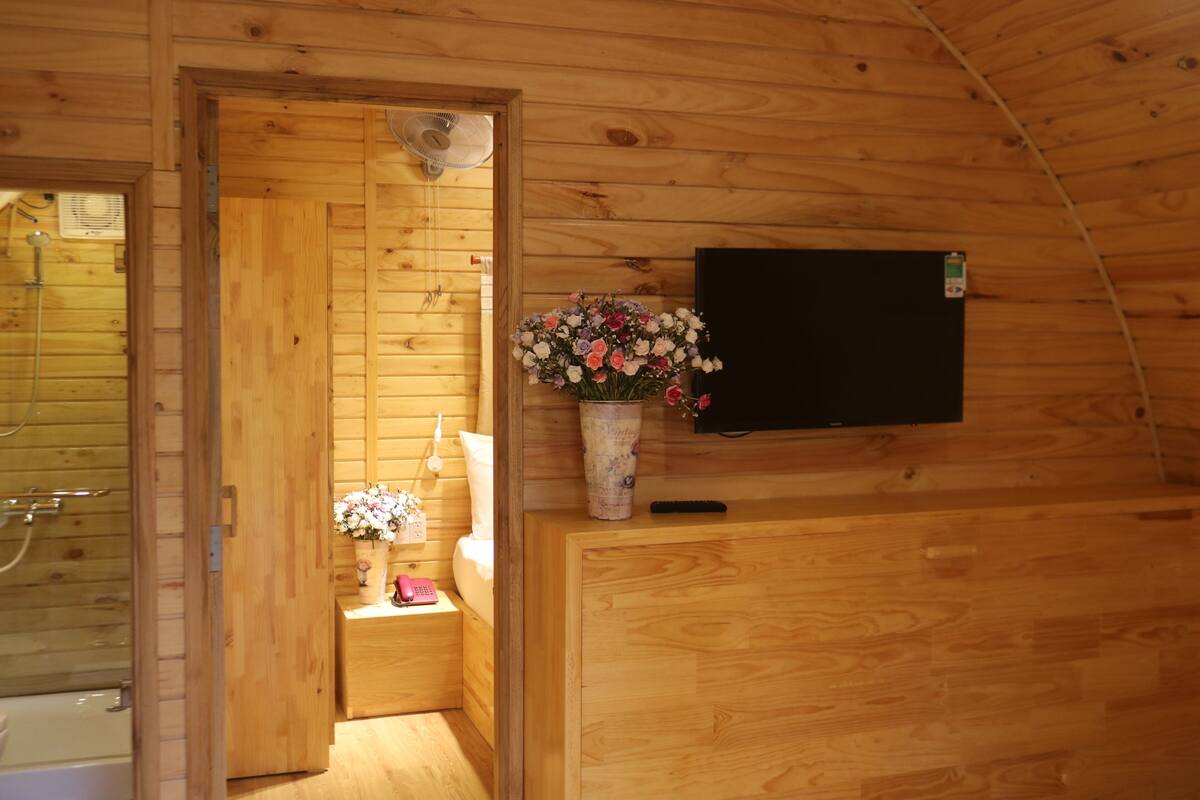 A wooden cabin interior with a wooden door, a small bathroom with a shower, a wooden cabinet, and a vase with flowers.
