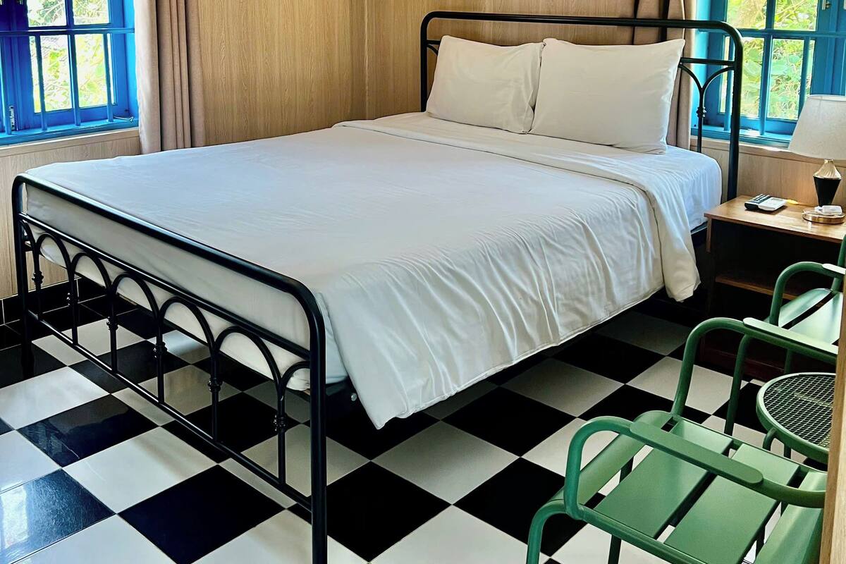 A room with a checkered floor, a bed with white bedding, a green chair, and a table with a lamp.