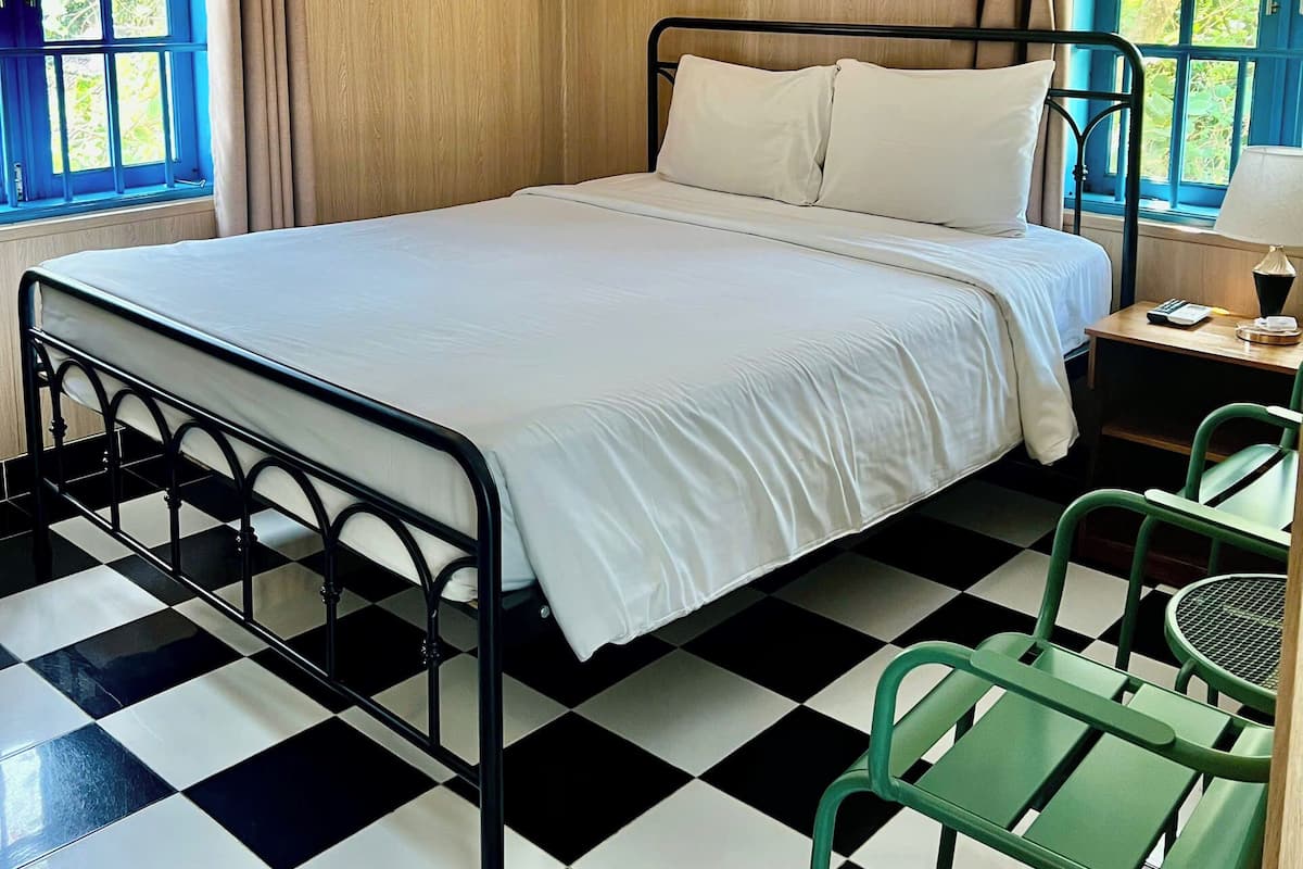 Una habitación con piso a cuadros, una cama con ropa de cama blanca, una silla verde y una mesa con una lámpara.