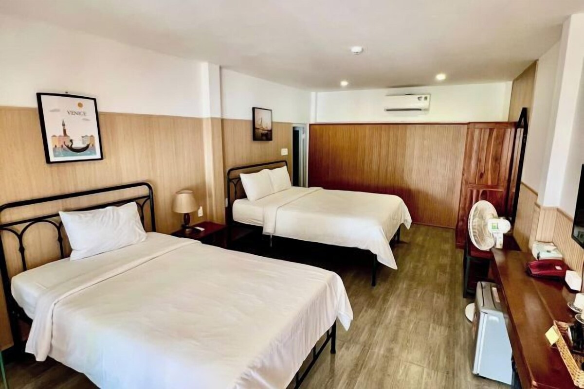 A hotel room with two beds, a desk, a chair, a fan, and a picture on the wall.