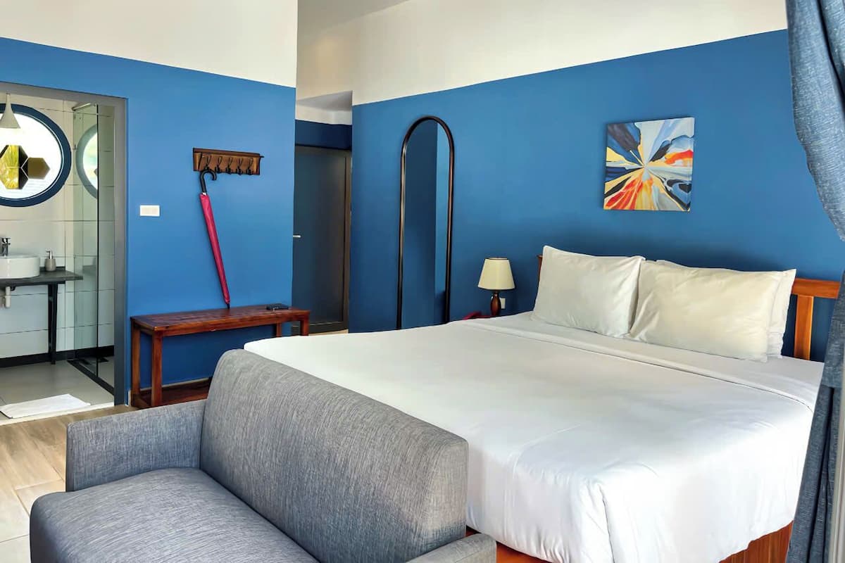 A hotel room with a bed, a bedside table, a lamp, a painting on the wall, and a bathroom visible through an open door.