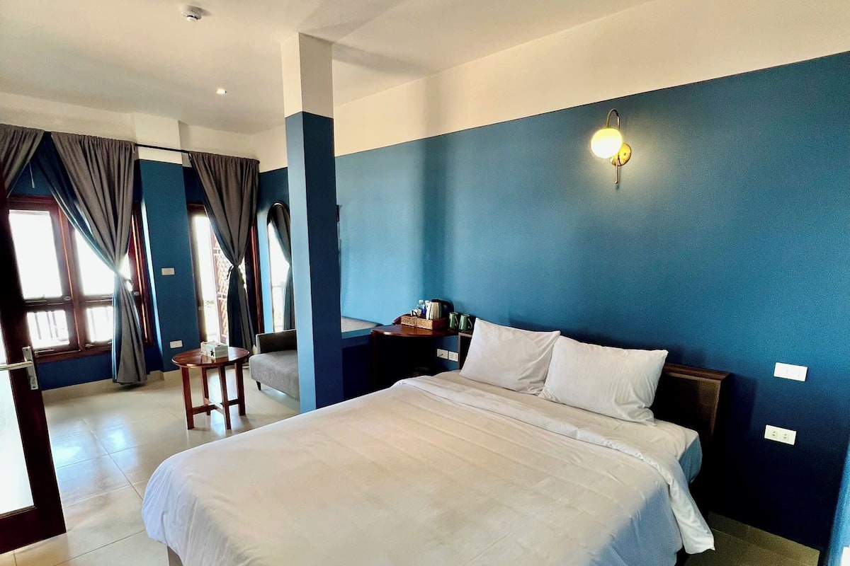 A hotel room with a large bed, a desk, and a chair. There is a window with curtains, a wall-mounted light, and a door.