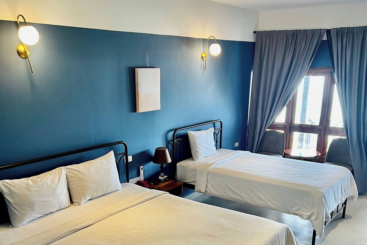 A hotel room with two beds, a bedside table with a lamp, a small table, and a window with curtains.