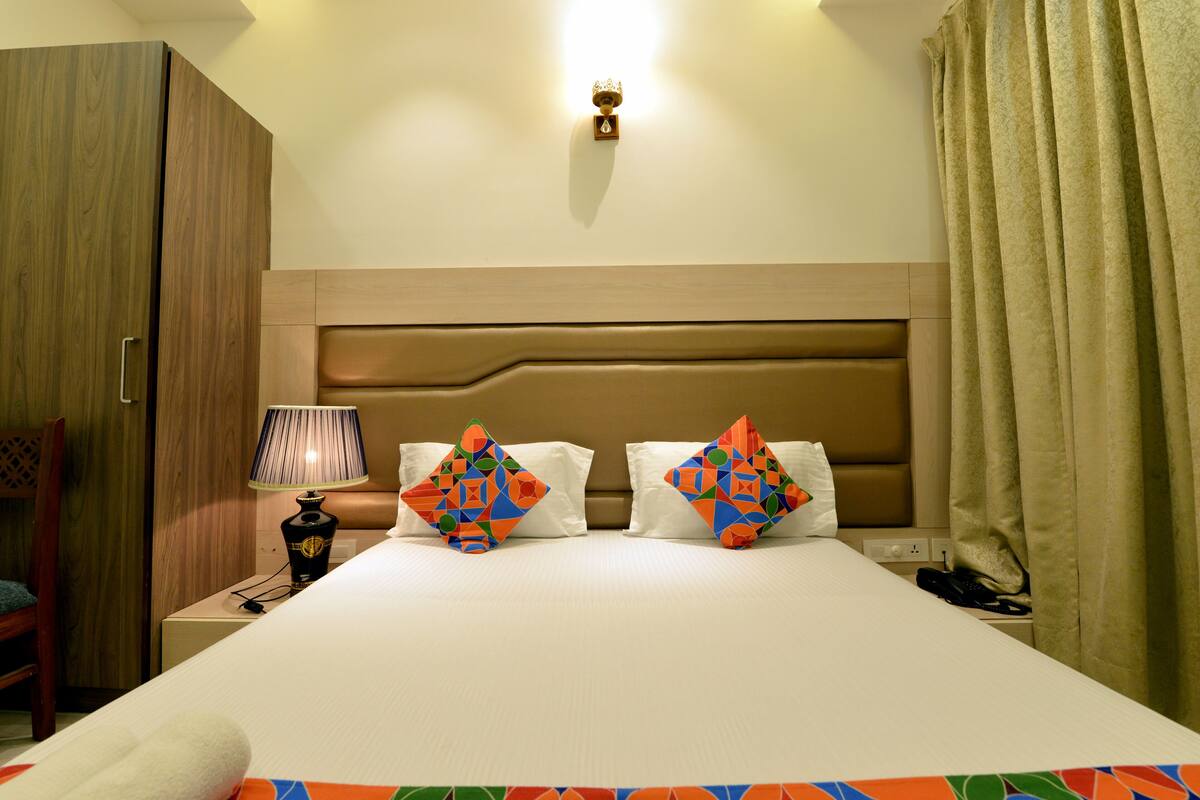 Una habitación de hotel con una cama grande, dos almohadas con diseños geométricos, una lámpara de noche, un armario de madera y una cortina.