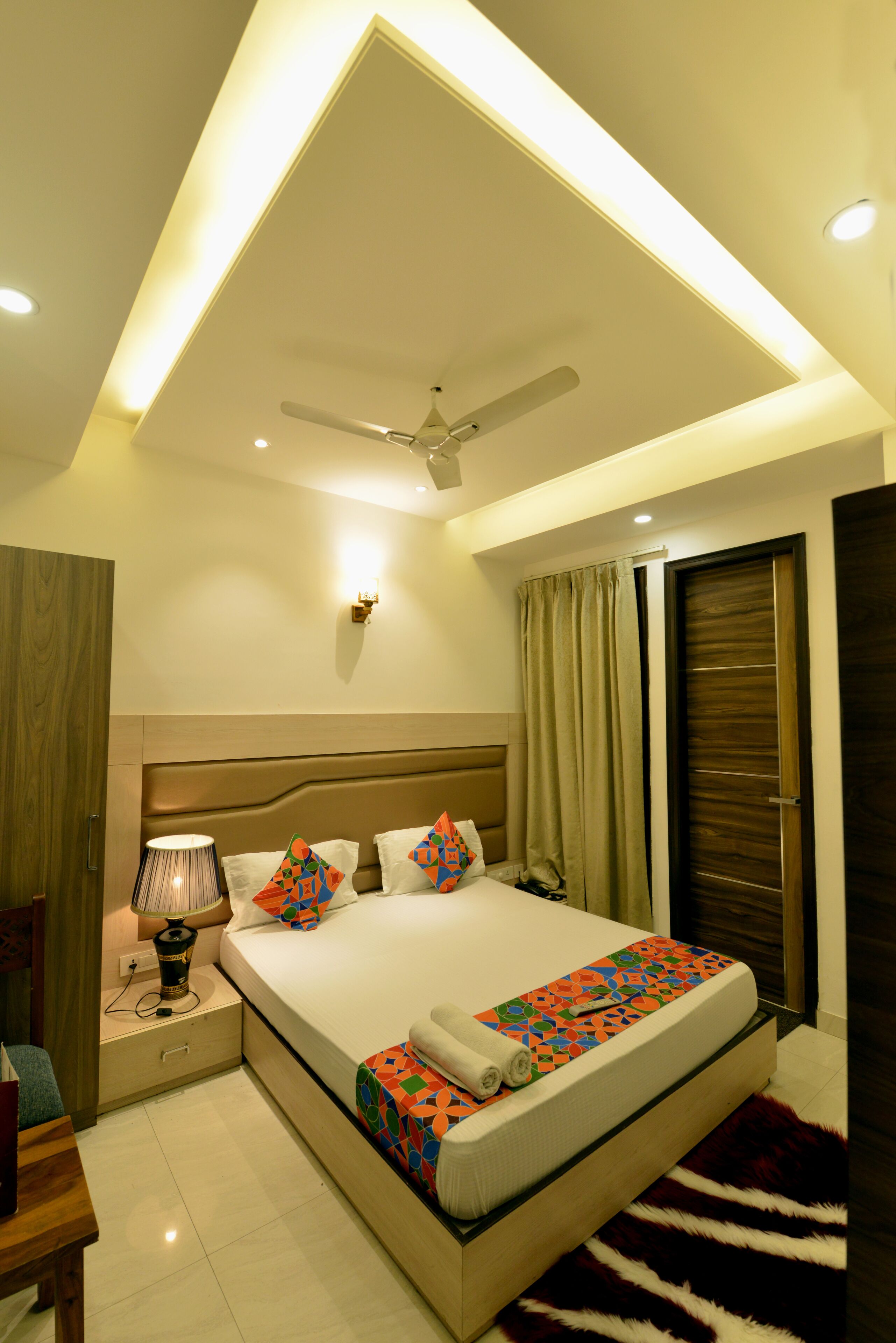 A modern hotel room with a large bed, a ceiling fan, a nightstand with a lamp, and a view of the outside through a window.