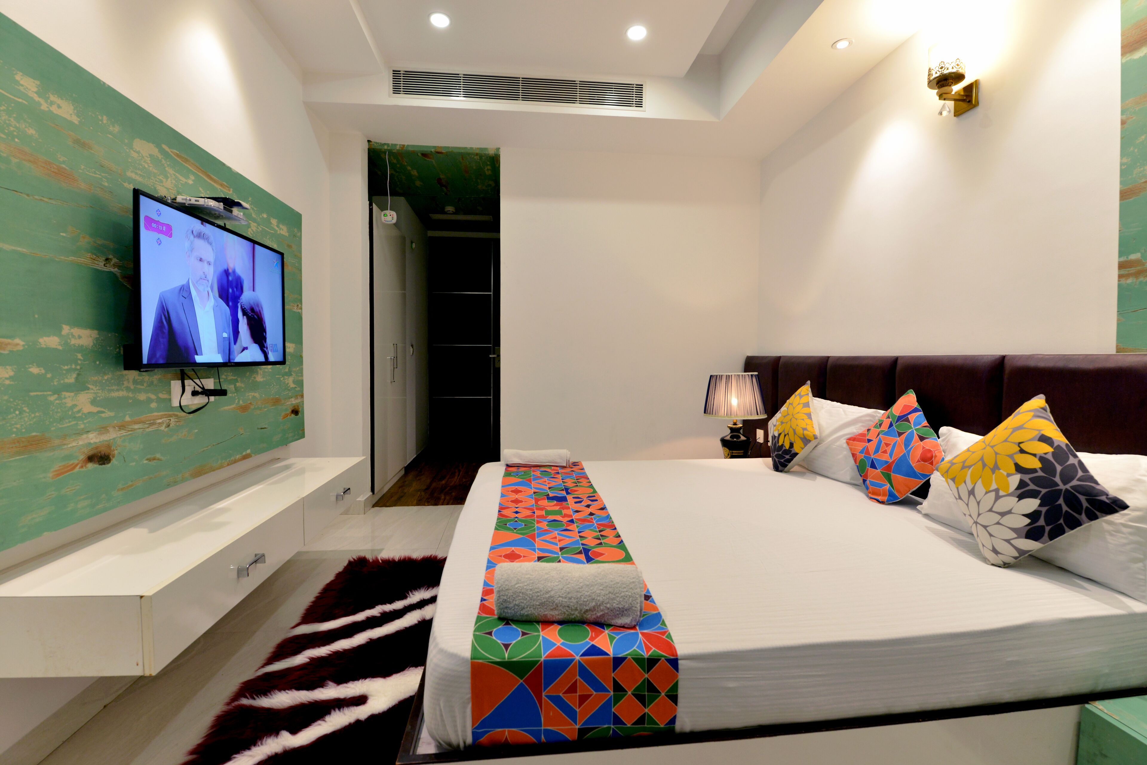 Una habitación de hotel moderna con una cama grande, almohadas coloridas, un televisor de pantalla plana montado en una pared verde, una mesita de noche blanca y un vestidor.