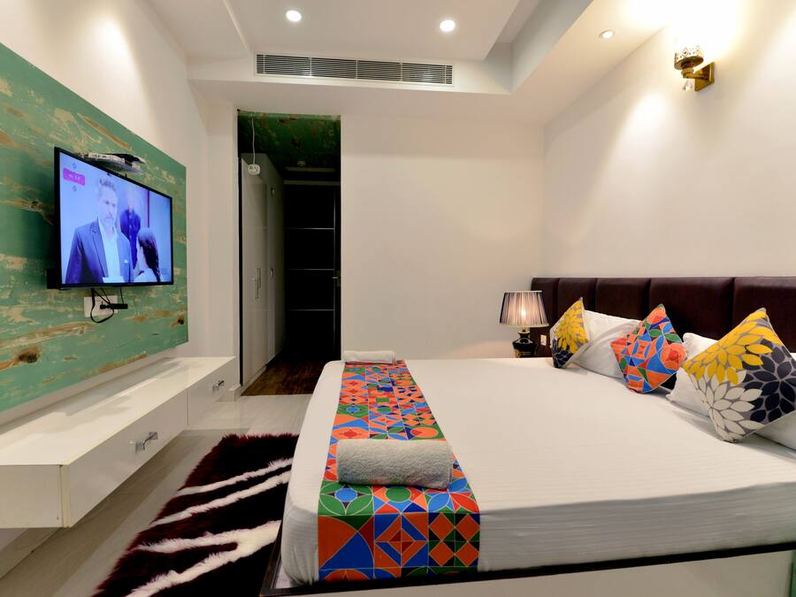 Una habitación de hotel moderna con una cama grande, almohadas coloridas, un televisor de pantalla plana montado en una pared verde, una mesita de noche blanca y un vestidor.