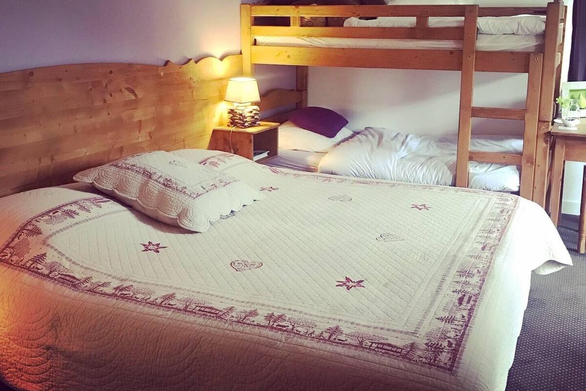 A bunk bed room with a wooden headboard, a bedside table with a lamp, a teddy bear, and a quilted bedspread.