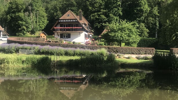 A house with a sloped roof, a balcony with red flowers, and a pond with a boat. - Chambres d'Hotes Maison Efftermatten (Andlau)