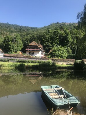 A house with a sloped roof, a balcony with red flowers, and a pond with a boat. - Chambres d'Hotes Maison Efftermatten (Andlau)