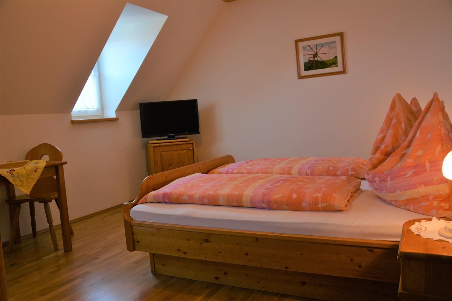 A bedroom with a sloped ceiling, a bed with patterned bedding, a television on a wooden cabinet, and a chair with a blanket.