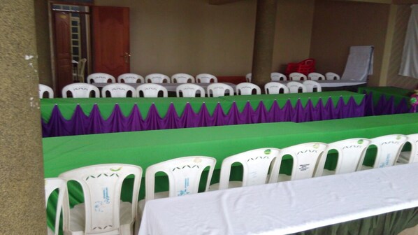 Un salón de conferencias con filas de sillas de plástico blanco, manteles verdes y morados, y una mesa blanca.