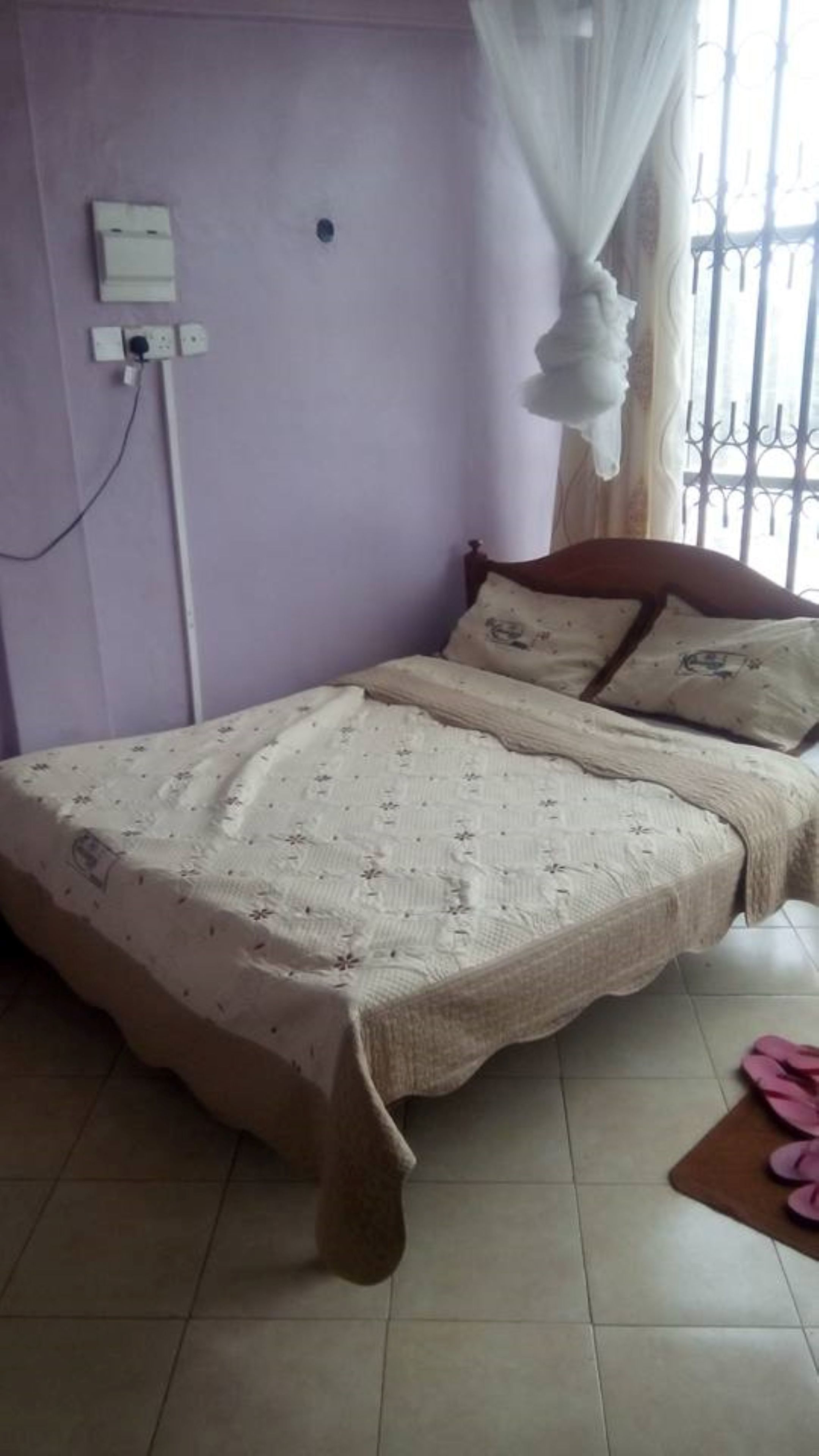 A bedroom with a bed, a power outlet, a window with bars, and a pair of pink slippers on the floor.