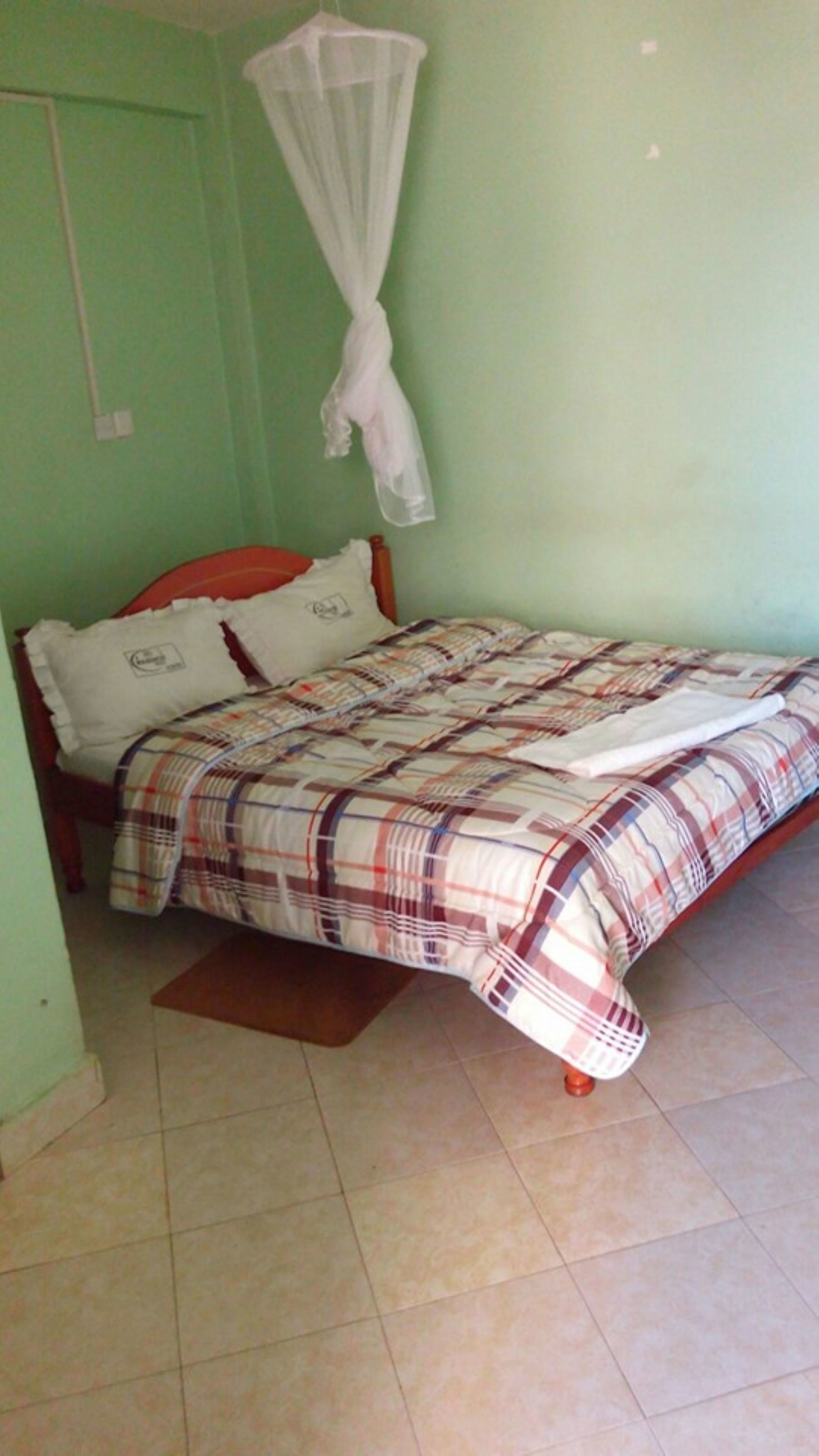 Una cama individual con una colcha escocesa y almohadas, una mosquitera sobre la cama y un suelo de baldosas.