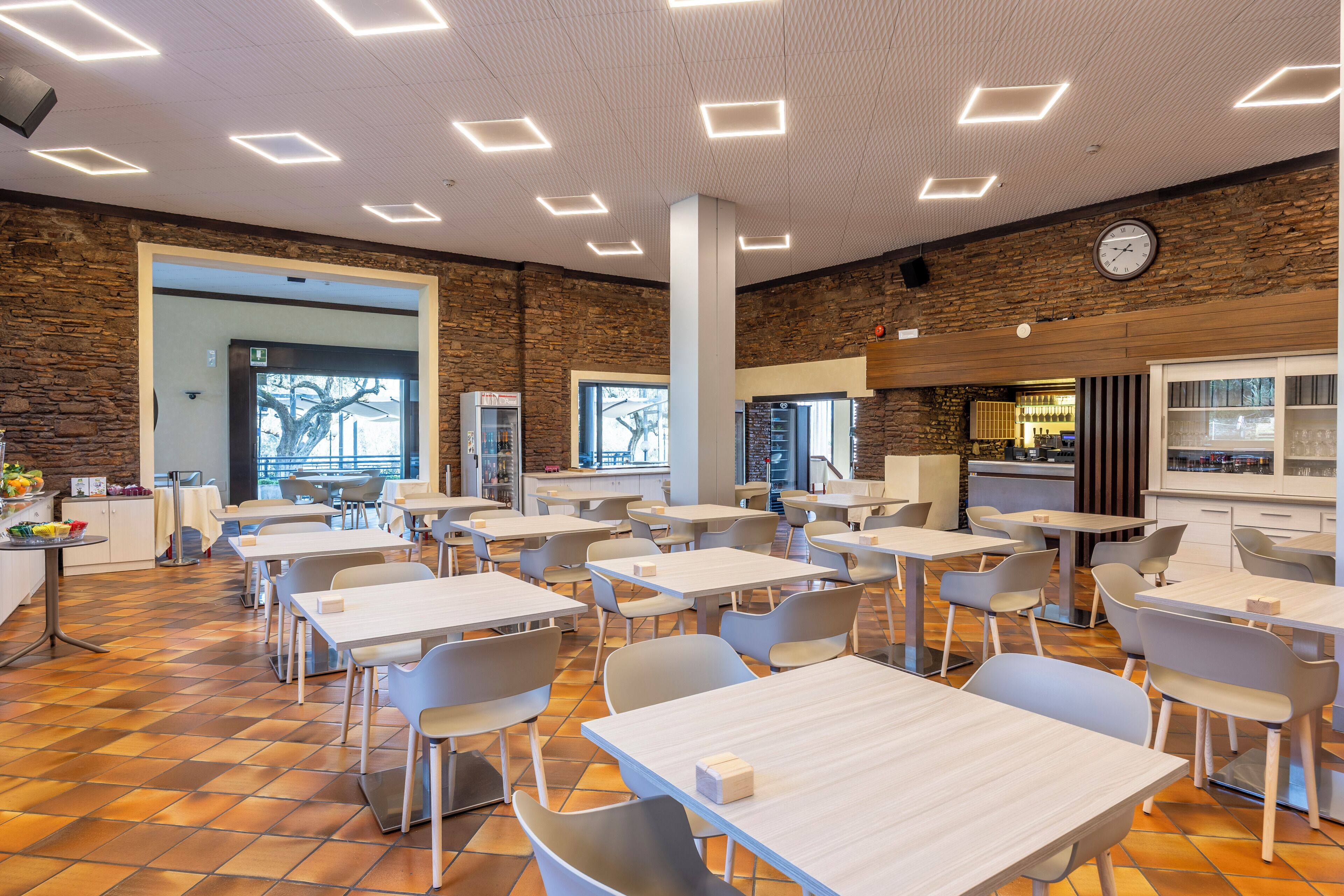 A spacious cafeteria with multiple tables and chairs, a clock on the wall, and a stone wall.