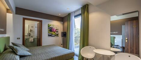 Una habitación de hotel moderna con cama, dos sillas, una mesita, un cuadro y un balcón con cortinas.