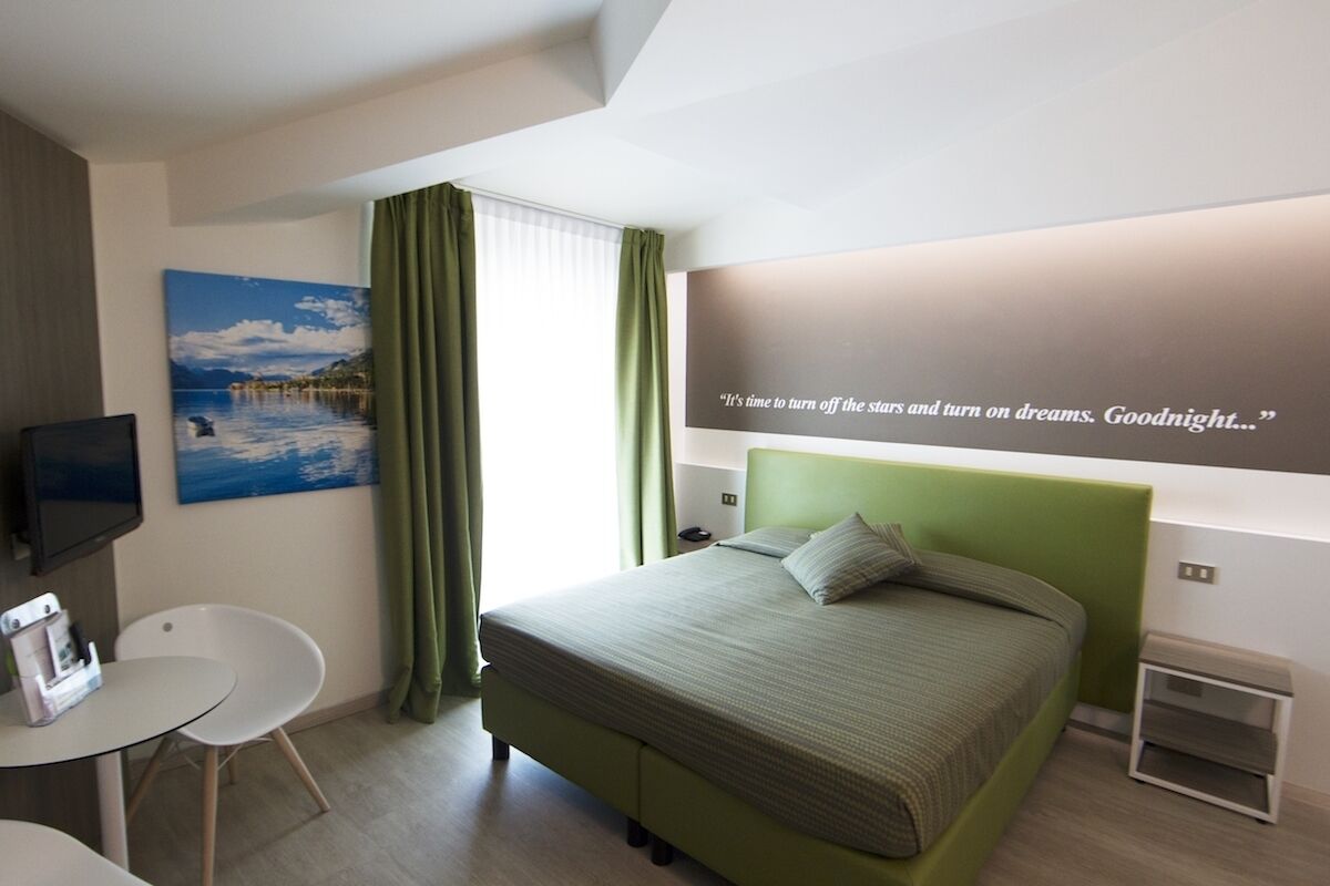 Una habitación de hotel moderna con una cama verde, un escritorio con una silla, un televisor de pantalla plana y un cuadro de una escena costera.