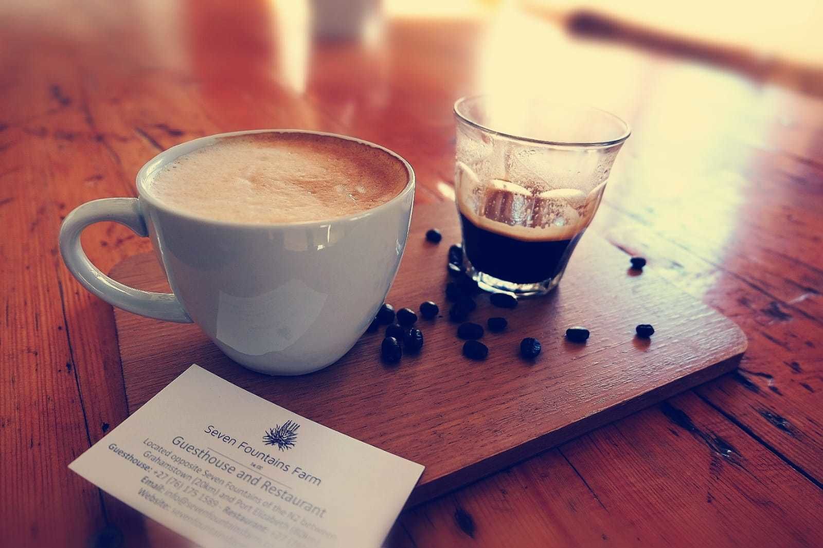 A white cup of coffee and a glass of dark liquid on a wooden table with scattered coffee beans. A business card for 'Seven Fountains Farm' is visible.