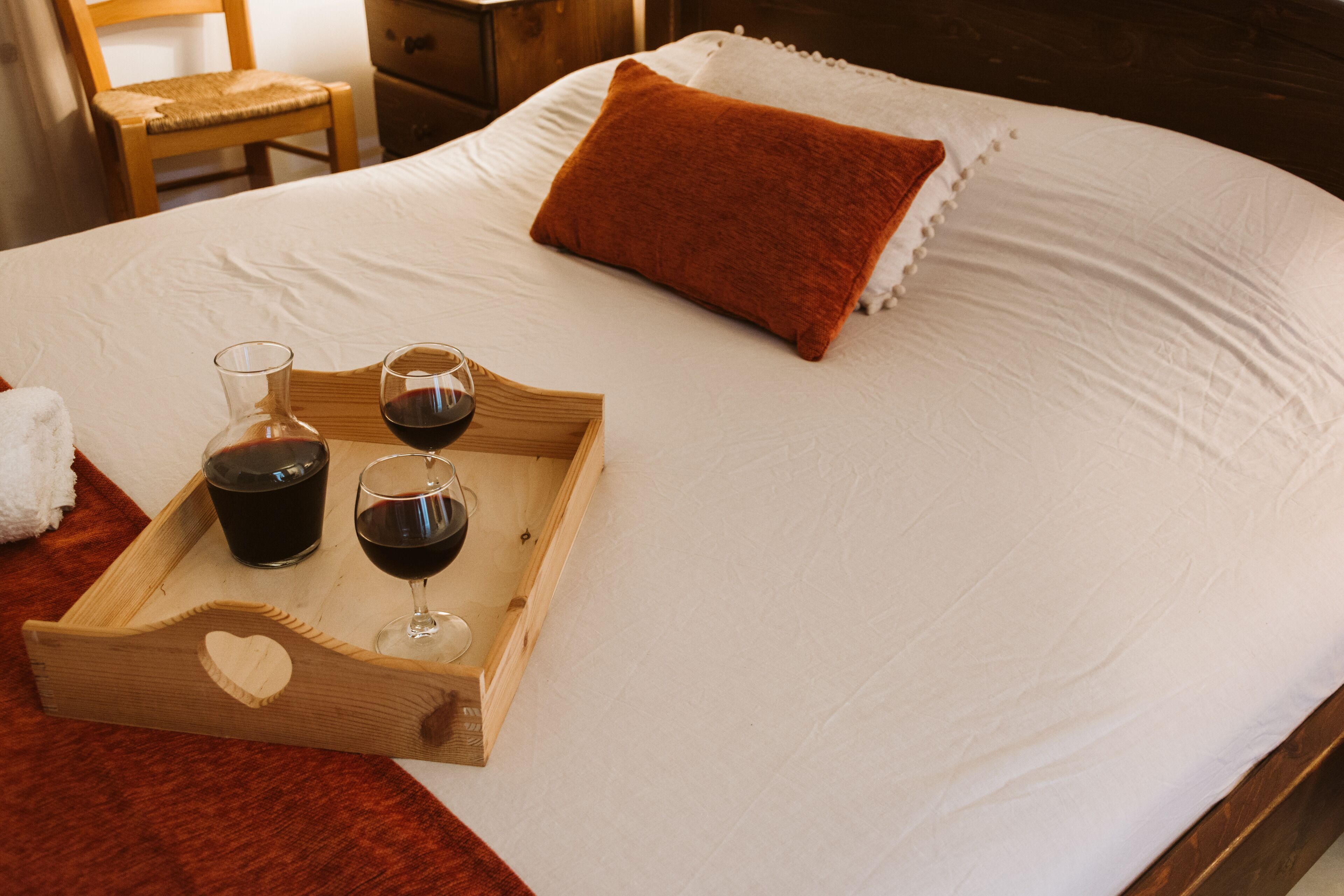 Cama bem arrumada com uma bandeja de madeira que contém dois copos de vinho tinto e uma garrafa, um travesseiro branco e um cobertor vermelho.
