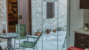 A room with a stone wall, a wooden ceiling, a glass door, a table with a red tablecloth, and a chair. - Ariadni Gaia Cottage (Limassol)