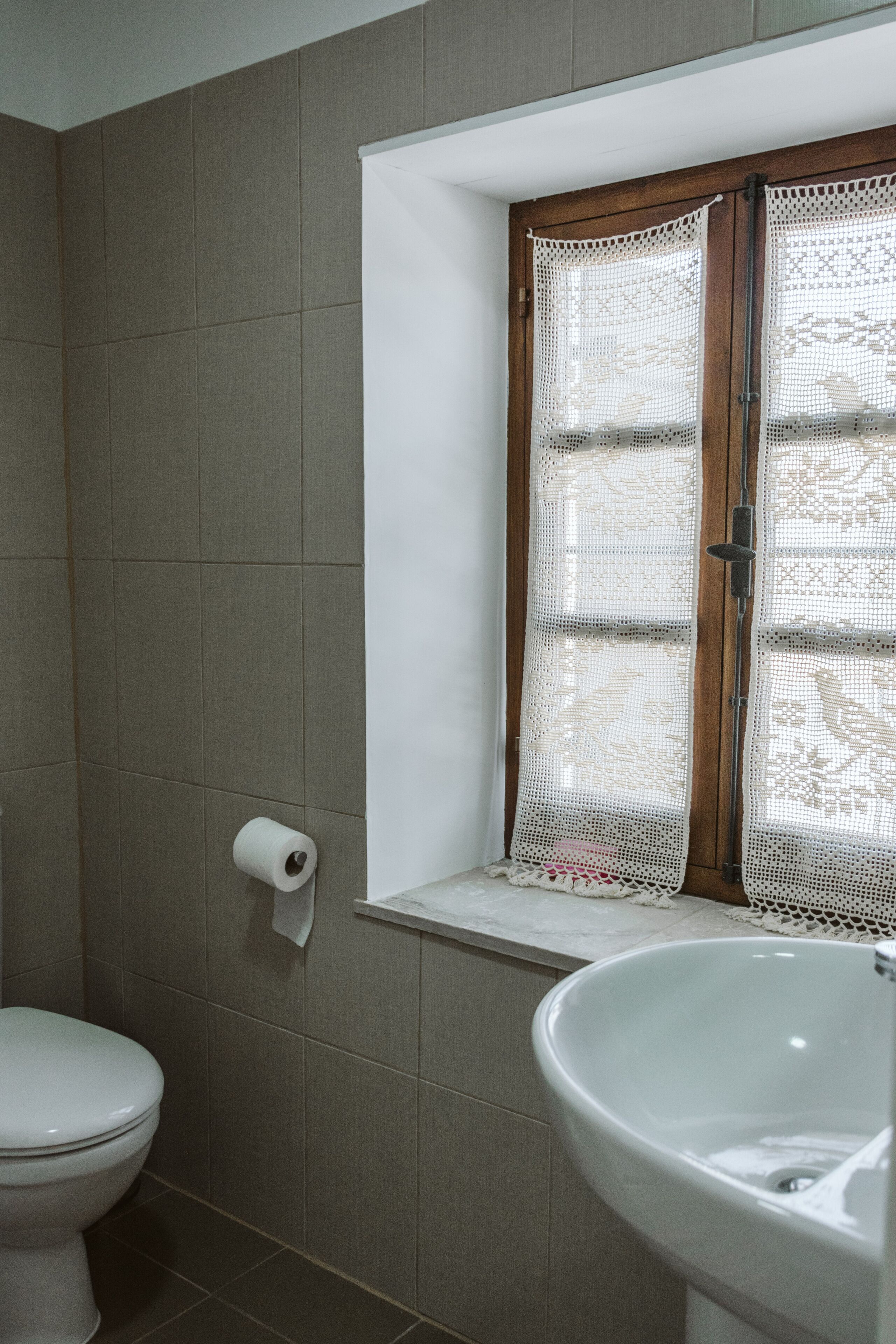 A bathroom with a toilet, a sink, and a window with lace curtains.