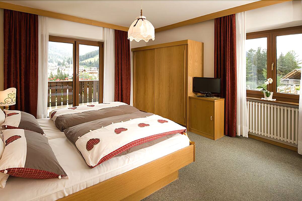 A bedroom with a large bed, wooden headboard, a television on a wooden cabinet, and a balcony with a view of the mountains.