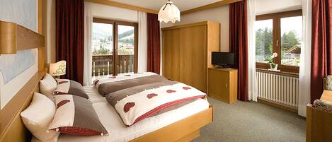 Un dormitorio con una cama grande, cabecera de madera, un televisor sobre un mueble de madera y un balcón con vistas a las montañas.