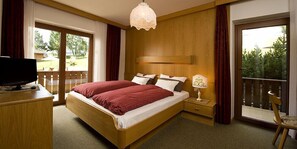 A bedroom with a bed, a TV on a wooden dresser, a nightstand with a lamp, and a balcony with a chair. - Mason Marmoleda (San Giovanni di Fassa)