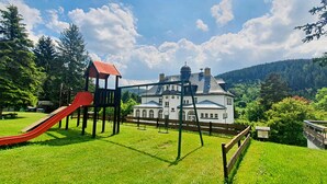 A playground with a red slide and swings, a white building with a red roof, and a forested hillside in the background. - Waldhotel Feldbachtal (Neuhaus am Rennweg)