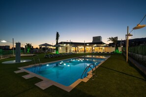 Una piscina con tumbonas y sombrillas, rodeada por un césped cuidado y un edificio con un letrero.