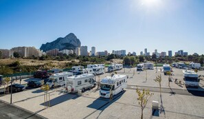 Un estacionamiento con vehículos recreativos y automóviles, rodeado por un paisaje urbano y una gran formación rocosa.