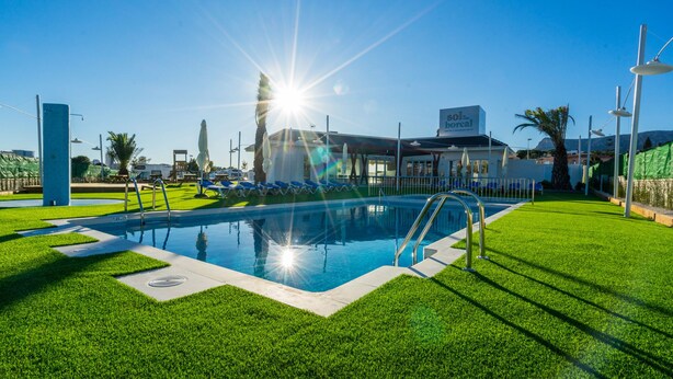 A swimming pool with a sunlit sky, a building with a sign, and green grass. - Camping Sol de Calpe Boreal - Caravan park (Calpe)